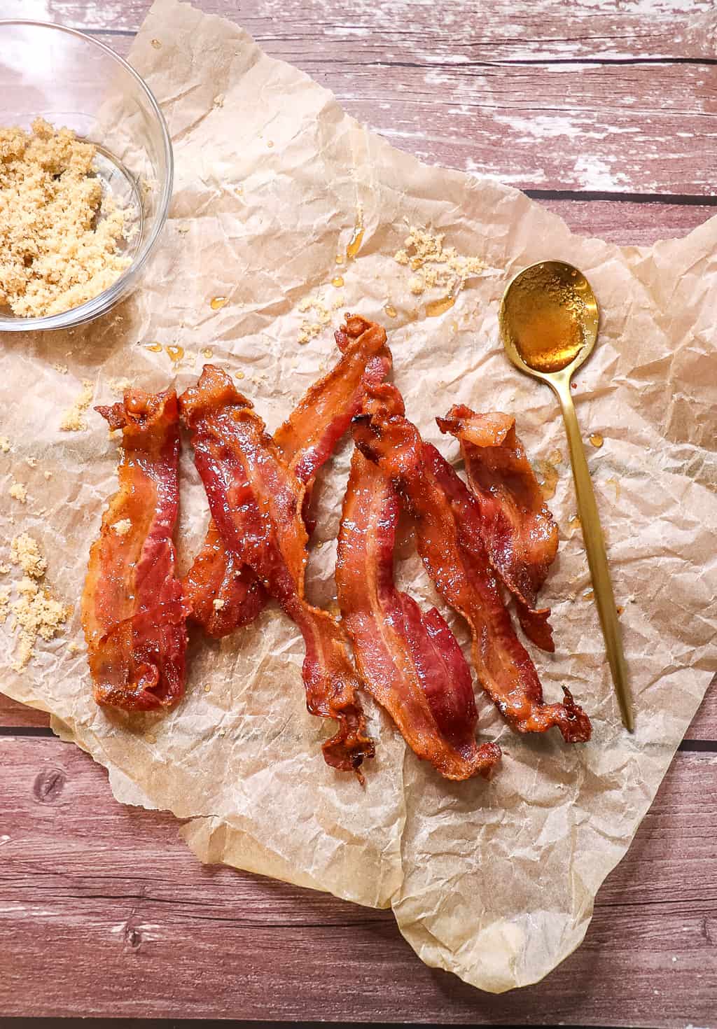 candied bacon on a piece of parchment paper with a spoon and a bowl of brown sugar next to it that are used as a prop