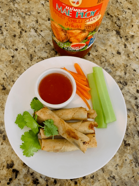 plated lumpia with sliced carrots, celery, cilantro, and a side of sweet chili sauce with the bottle in the background