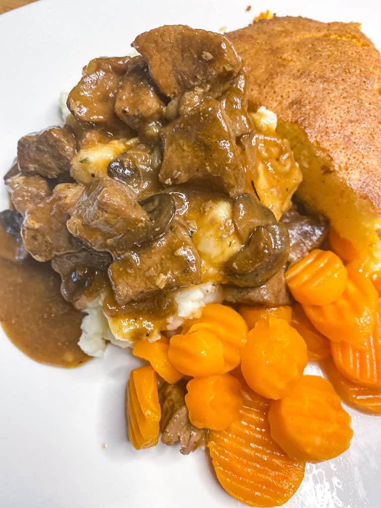 beef tips and gravy over mashed potatoes on a white plate with a side of carrots and cornbread