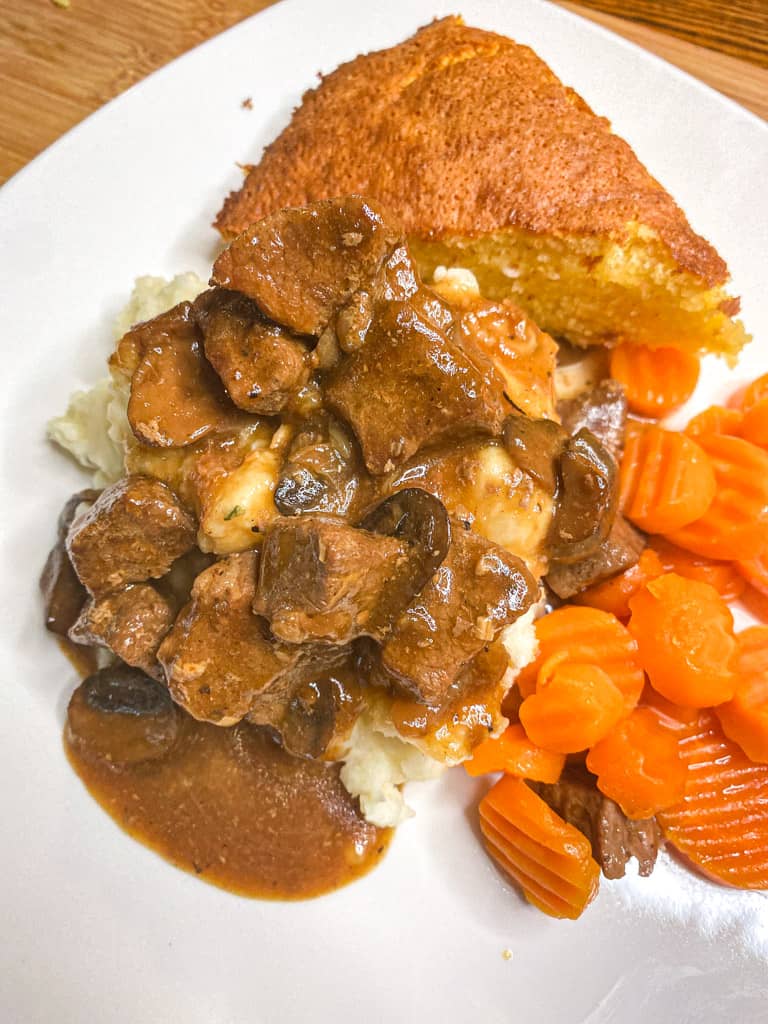 beef tips and gravy over mashed potatoes on a white plate with a side of carrots and cornbread