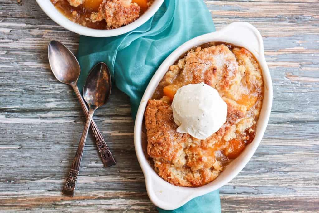 overhead view of a pan of peach cobbler topped with a large scoop of vanilla ice cream with a couple of spoons to the side