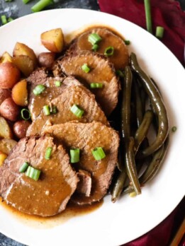 sliced pot roast on a plate with brown gravy and a side of roasted potatoes