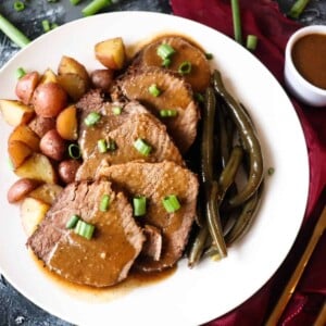 sliced pot roast on a plate with brown gravy and a side of roasted potatoes