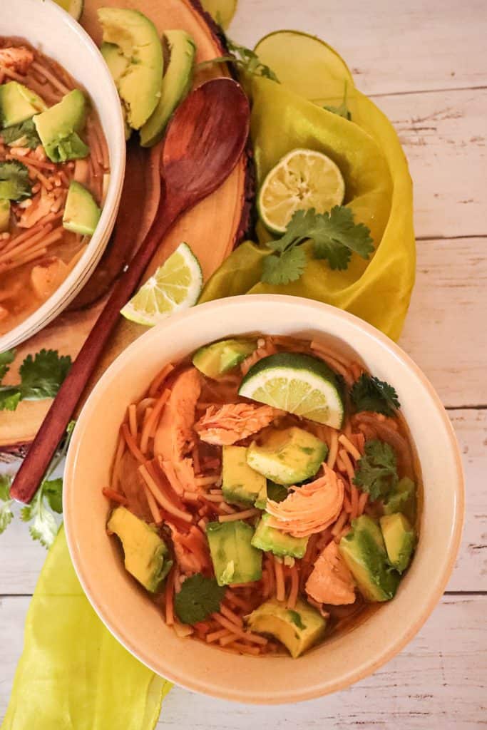chicken avocado lime soup A bowl of chicken avocado lime soup next to a large wooden bowl, sliced limes and avocados