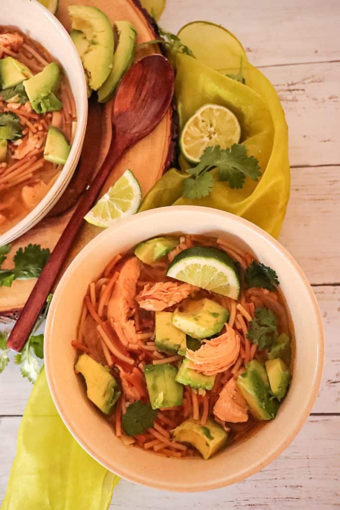 A bowl of chicken avocado lime soup next to a large wooden bowl, sliced limes and avocados