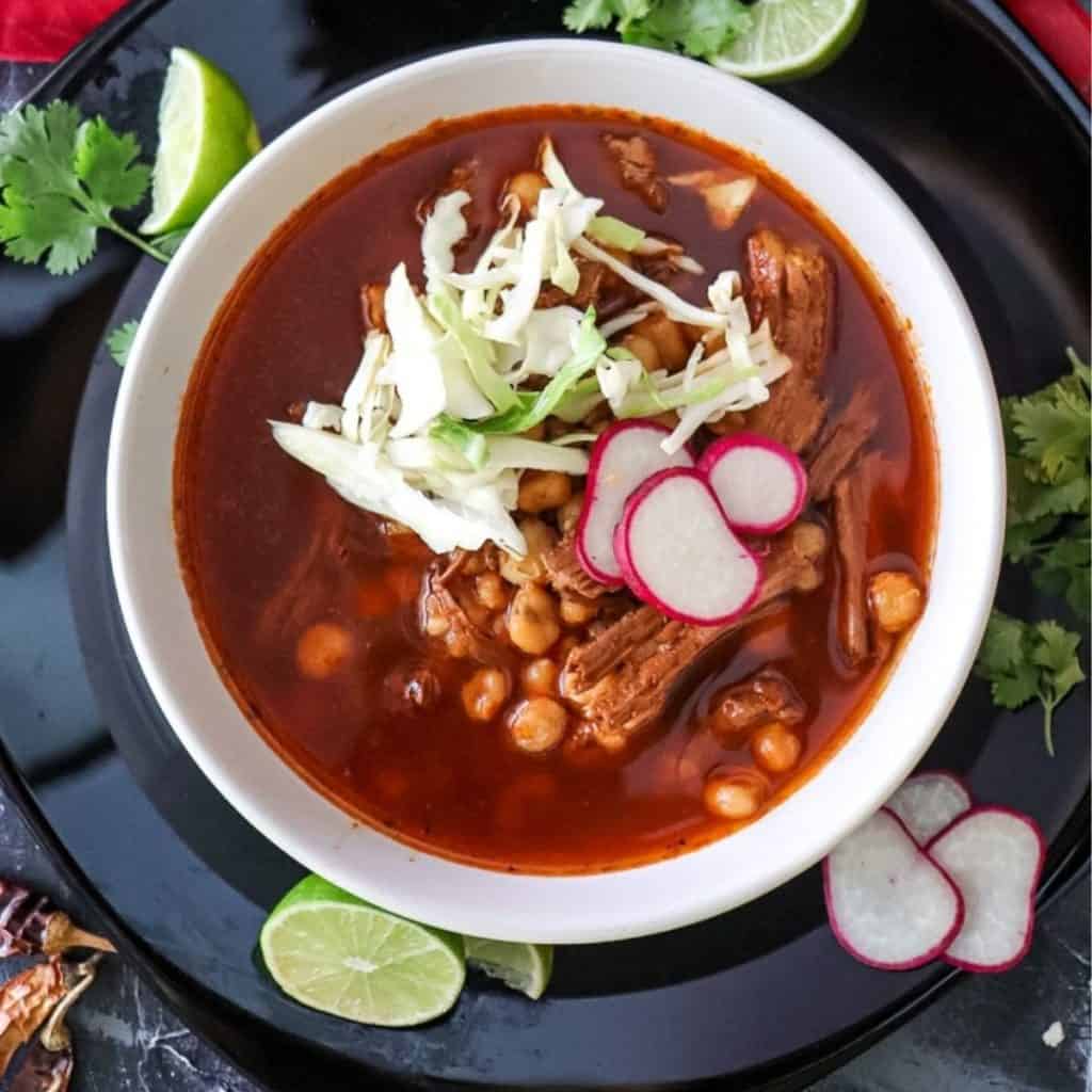 Overhead view of a bowl of Pozole Rojo with garnishes including cabbage, radishes and sliced limes.