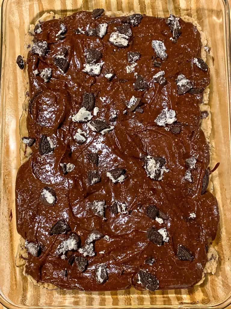 Brownie batter and chunks of Oreos placed over Oreos and chocolate chip cookie dough in a baking dish.