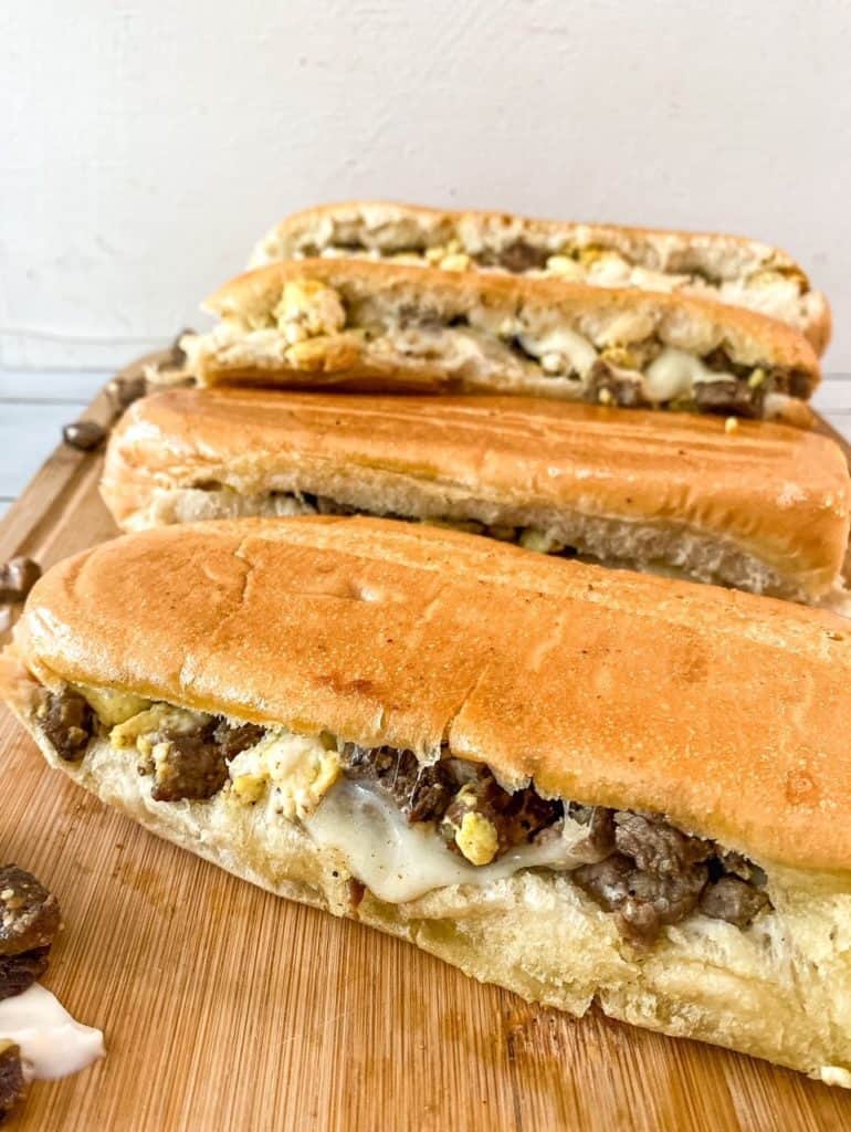 Four Steak, Egg and Cheese Breakfast Sandwiches lined up on a wooden cutting board.