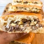 A hand holding a Steak, Egg and Cheese Breakfast Sandwich with more sandwiches in background on a wooden cutting board.