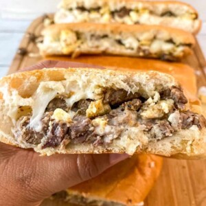 A hand holding a Steak, Egg and Cheese Breakfast Sandwich with more sandwiches in background on a wooden cutting board.