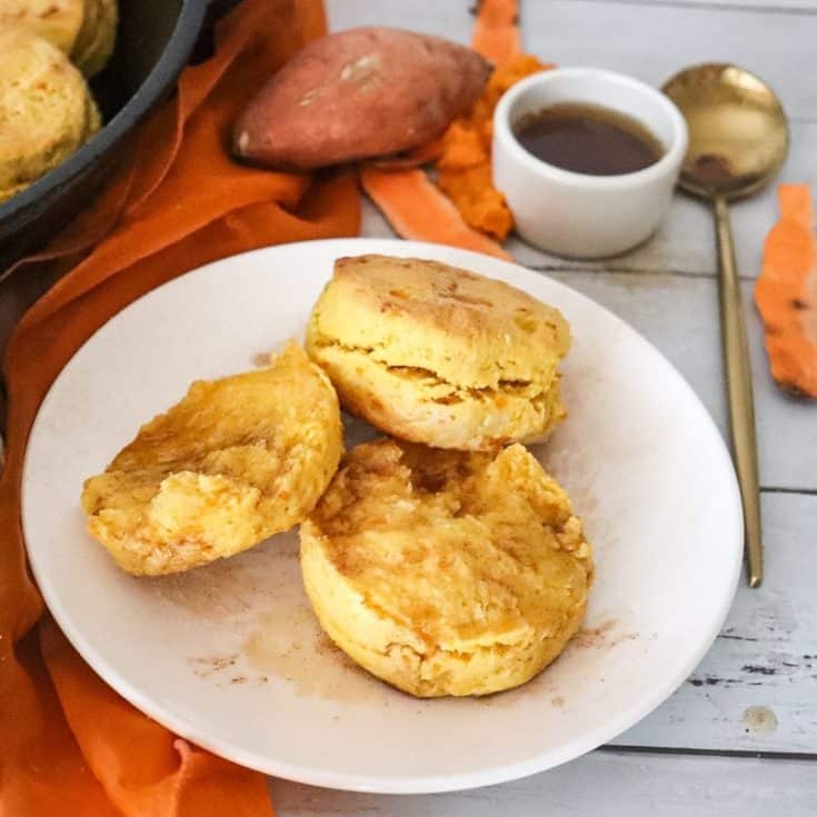 Two plated Cinnamon Sugar Sweet Potato Biscuits. One is sliced open and the other is closed. A sweet potato and a small bowl of cinnamon sugar butter are in the background.