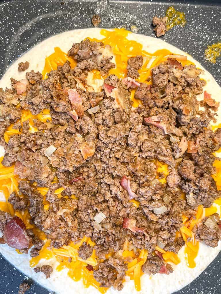 ground beef and bacon with cheese on top of a tortilla in a skillet for a cheeseburger quesadilla