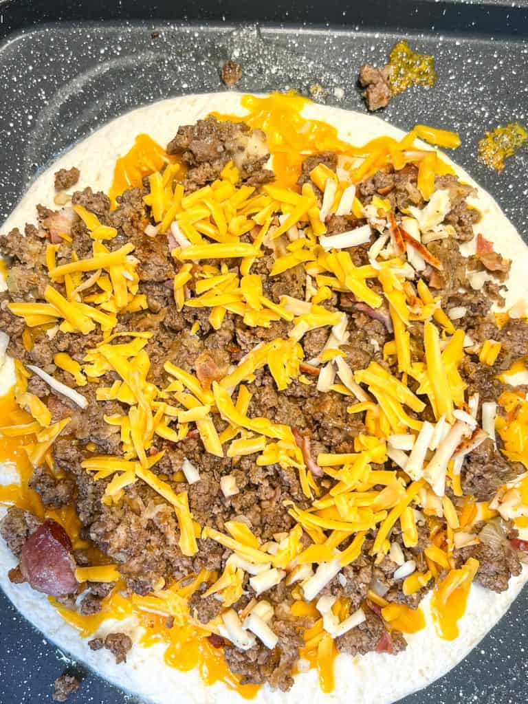 ground beef and bacon with cheese on top of a tortilla in a skillet for a cheeseburger quesadilla