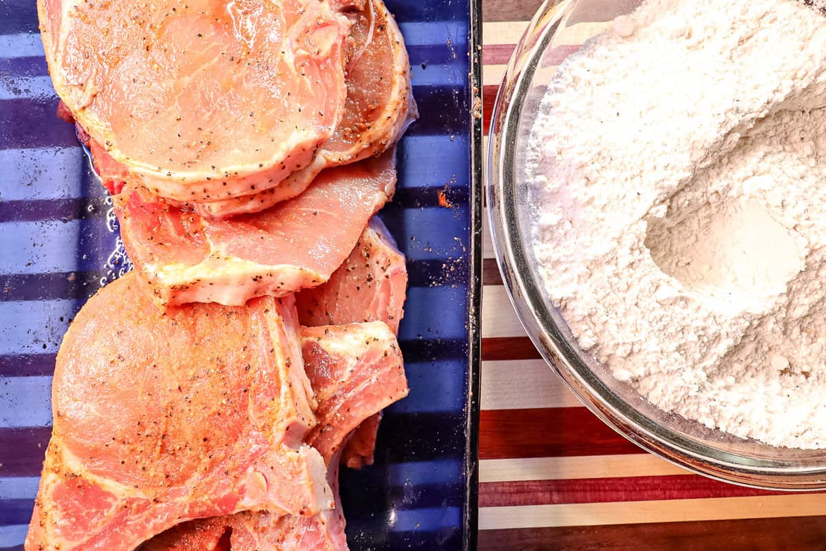 cleaned and seasoned raw pork chops are in one dish and seasoned flour is in another dish right beside it to bread