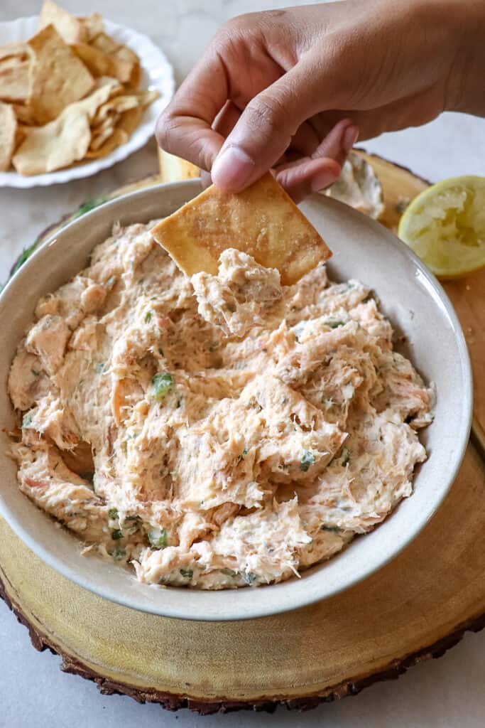 A hand holding a pita chip with smoked salmon dip on the tip directly over a bowl of dip.