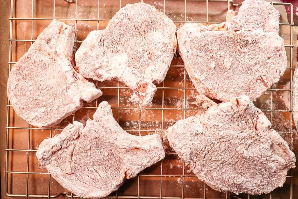 pork chops that have been covered in a seasoned flour breading are sitting on a cooling rack so that the breading can set