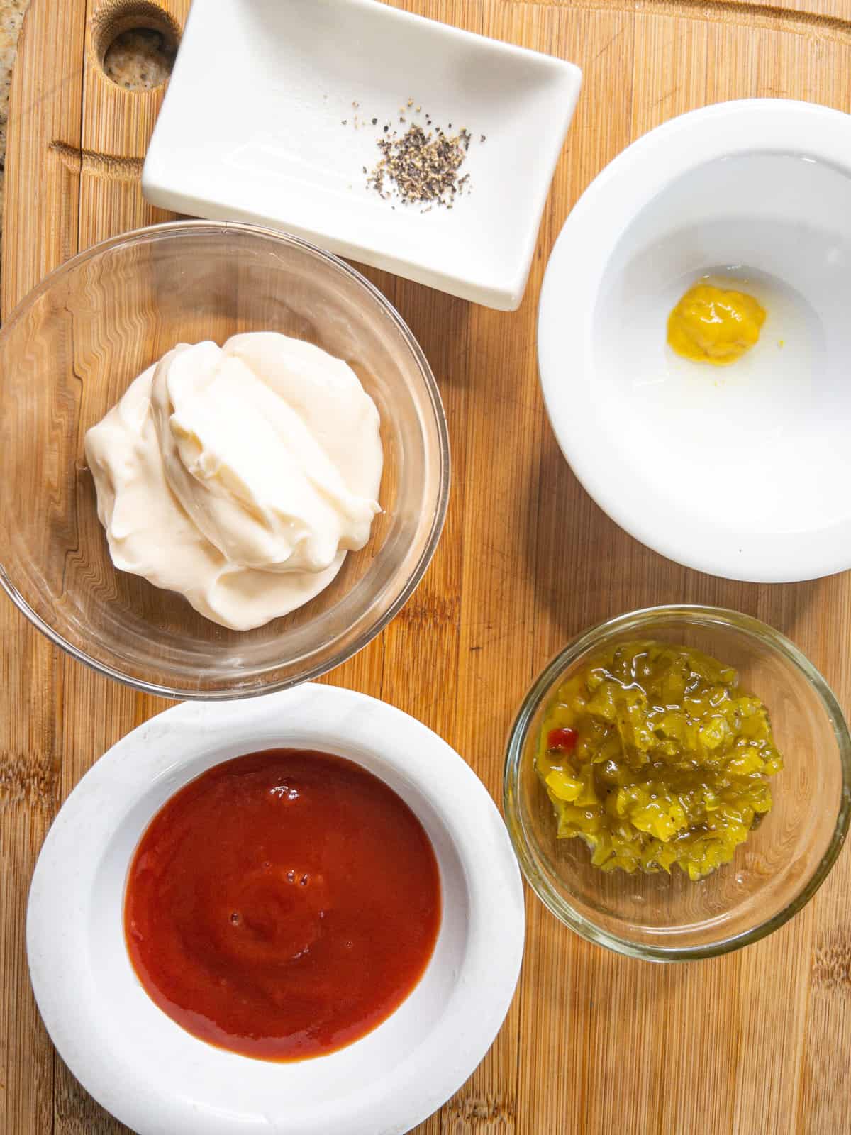 mayo, mustard, ketchup, sweet pickle relish, and salt and pepper in bowls on a cutting board