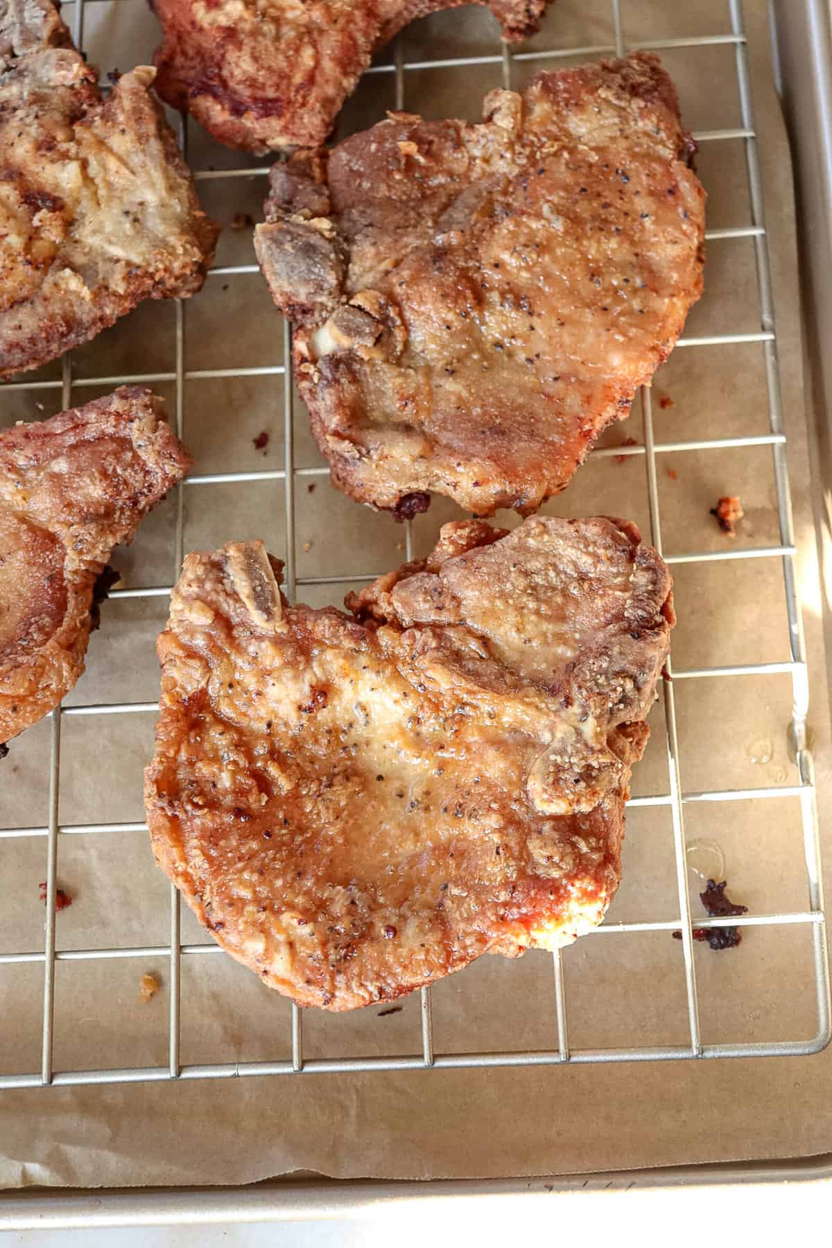 pork chops are resting on a cooling rack that is sitting over a piece of parchment paper so that all of the excess oil from frying can drain
