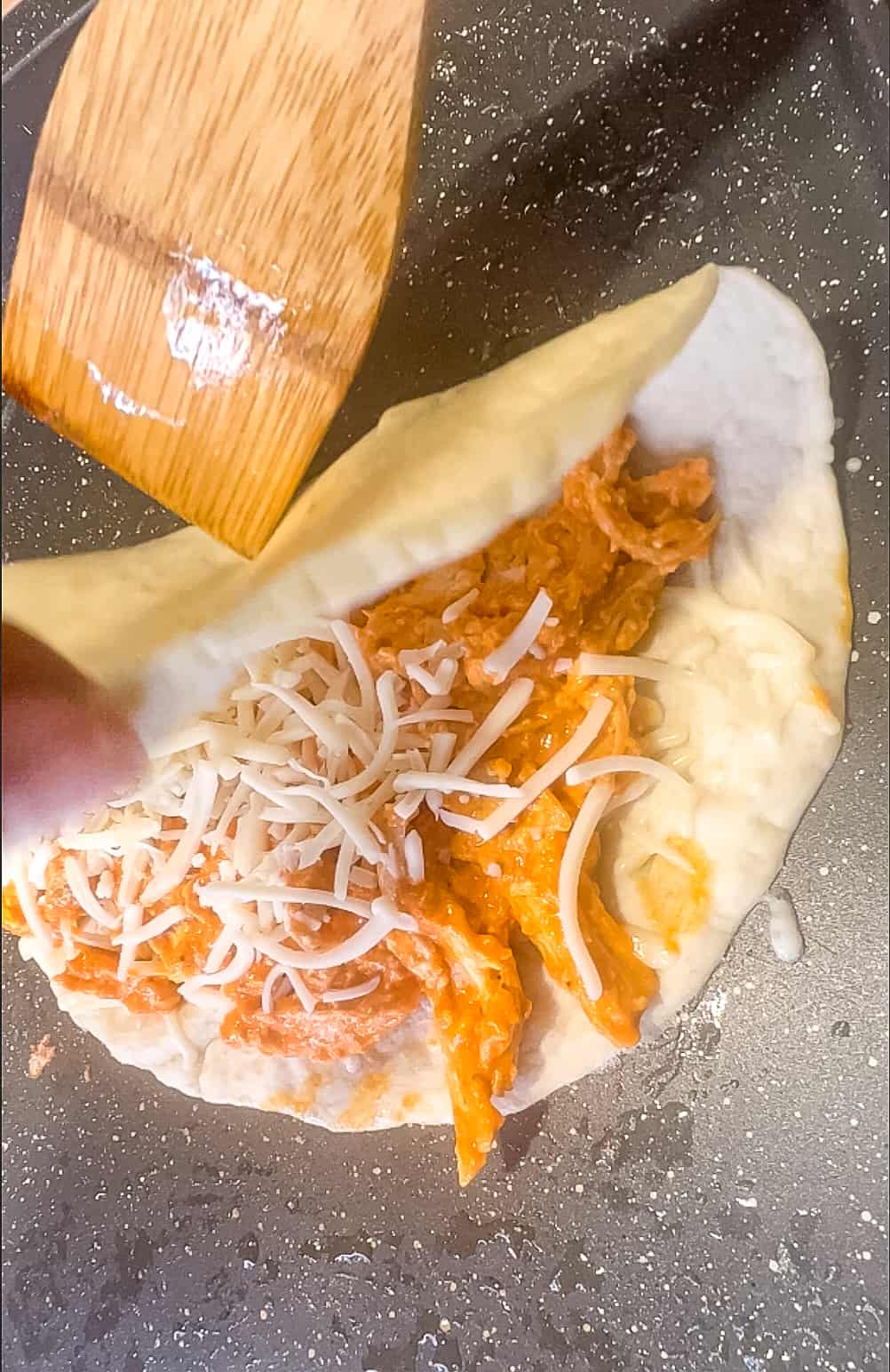 A tortilla filled with buffalo chicken mixture and cheese being folded in a skillet.