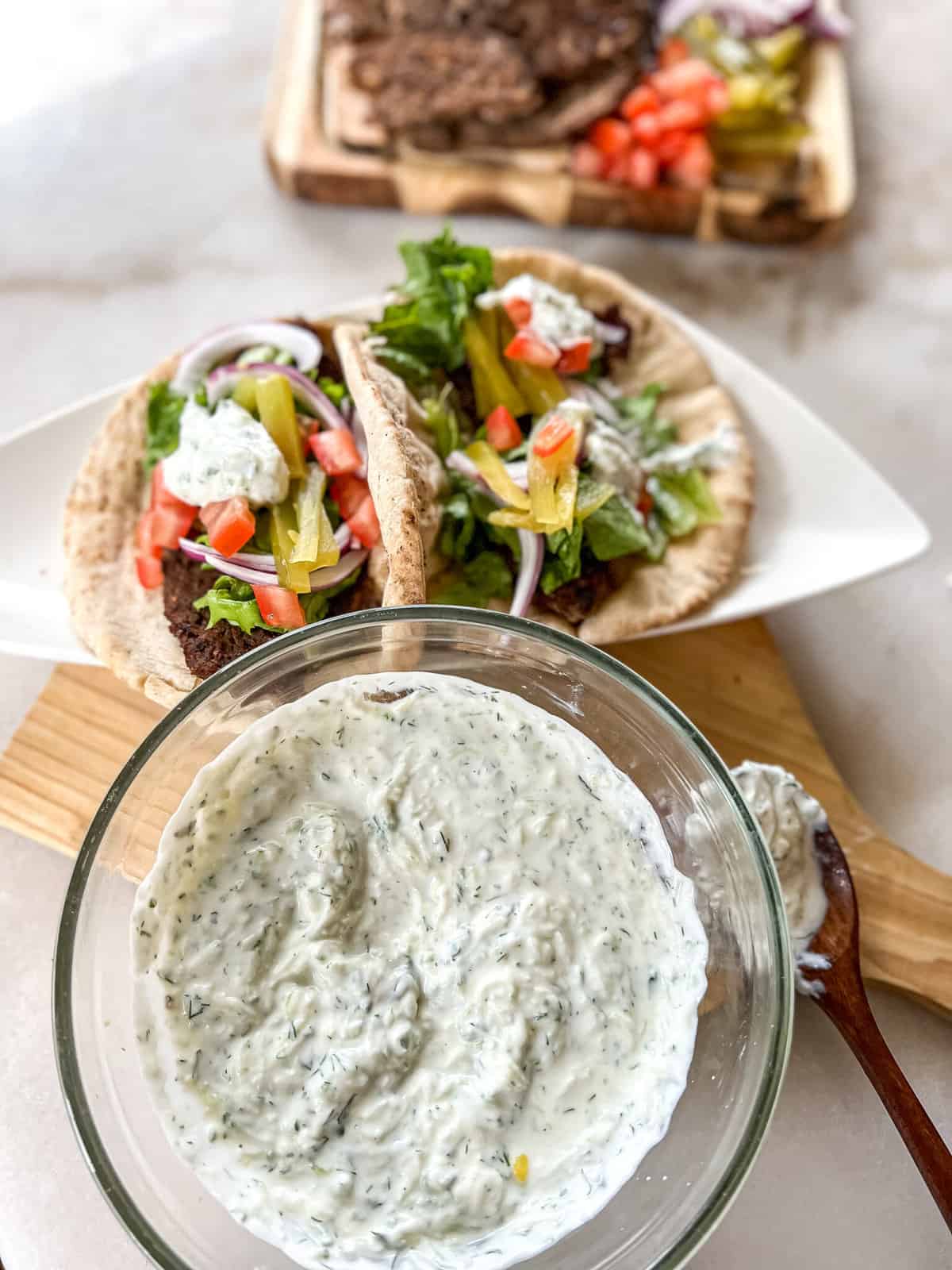 tzatziki sauce in a bowl with gyro sandwiches and gyro meat in the background