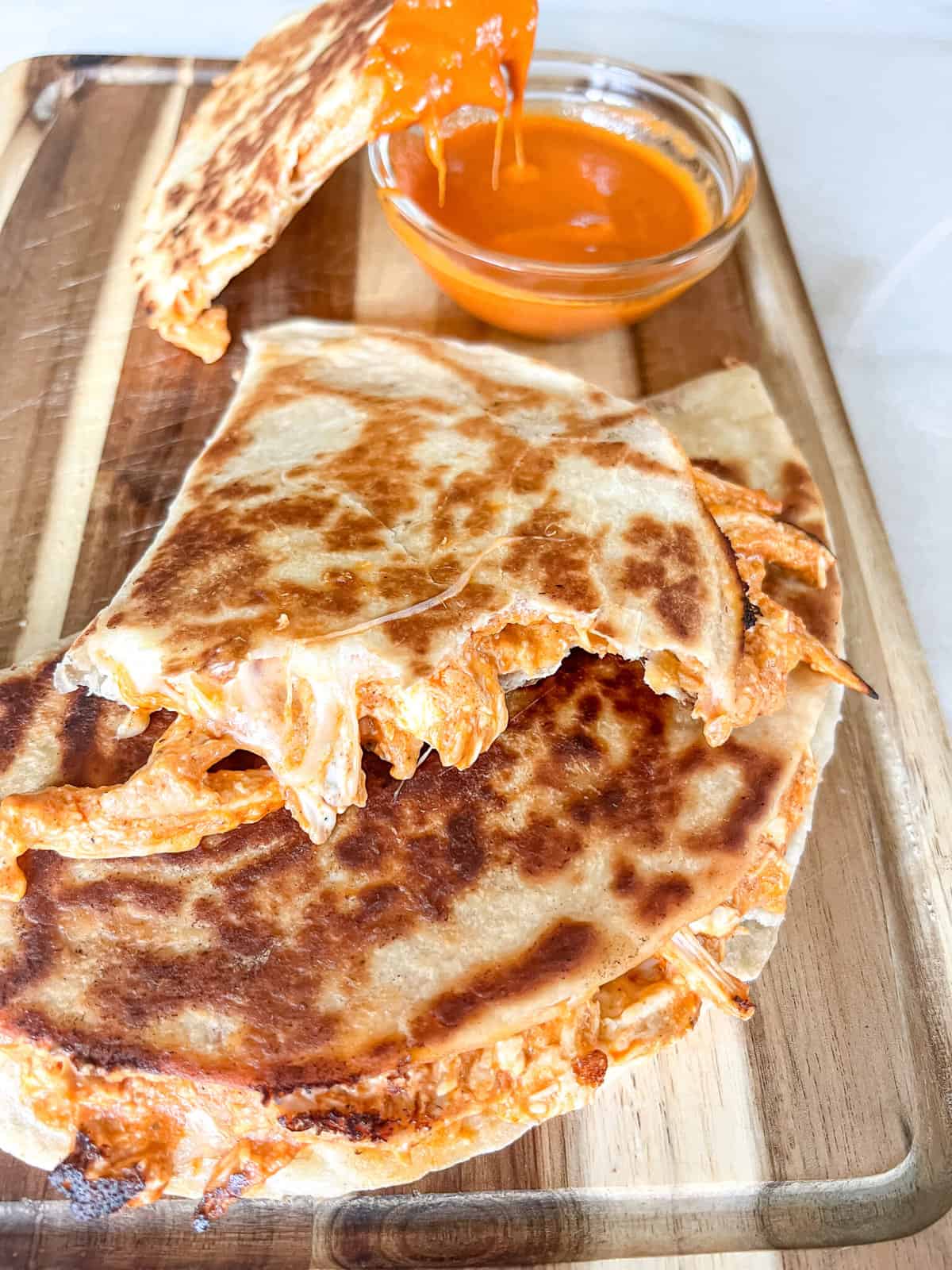 Platter of crispy quesadillas with a quesadilla dipped in buffalo sauce in the background.