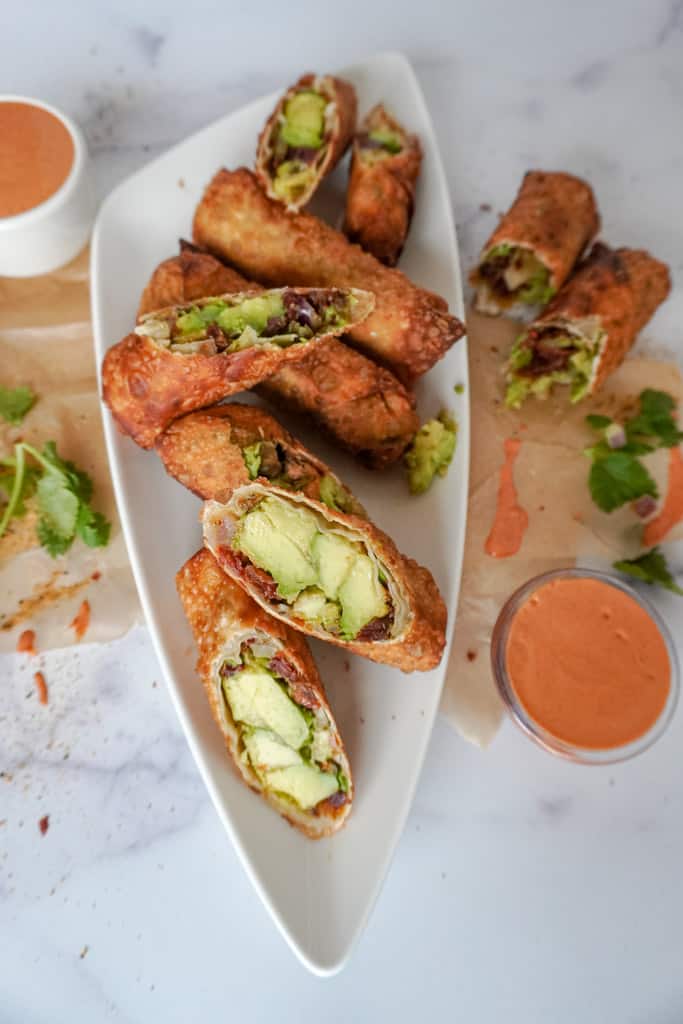 overhead view of a hand holding an egg roll over a platter of avocado egg rolls with sauce