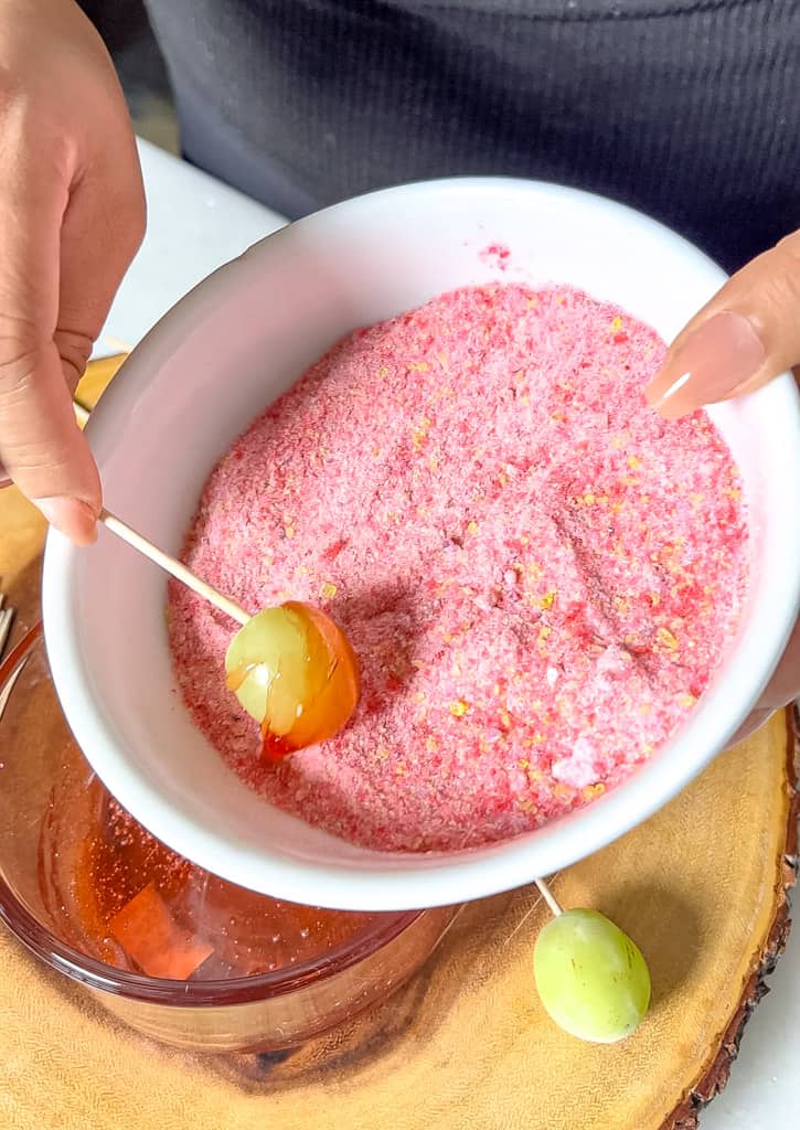 a single grape on a toothpick being dipped into a crushed candy mixture