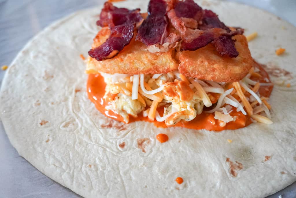 close up view of a tortilla with sauce, scrambled eggs, cheese, hash browns and bacon at the center