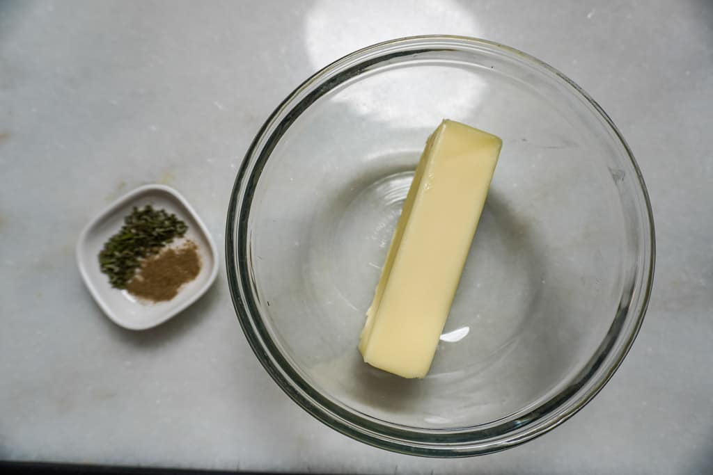 a bowl containing a stick of butter beside a small dish of dried herbs