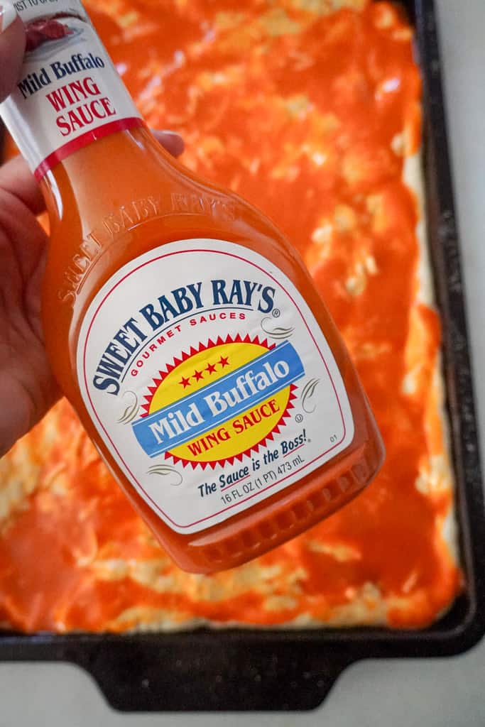 a hand holding a bottle of Sweet baby Ray's buffalo sauce over pizza dough on a baking sheet slathered with mild buffalo sauce
