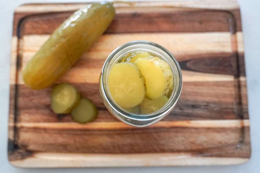 a whole pickle on a cutting board and sliced pickles in a jar next to it