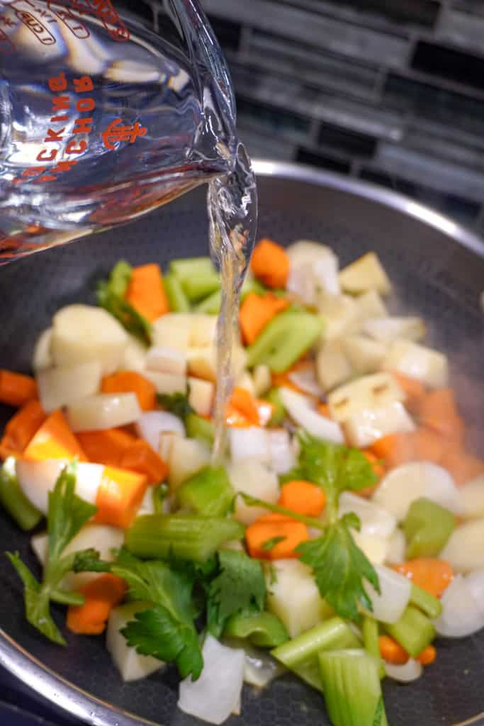 water being poured from a measuring cup into a skillet filled with sautéed veggies