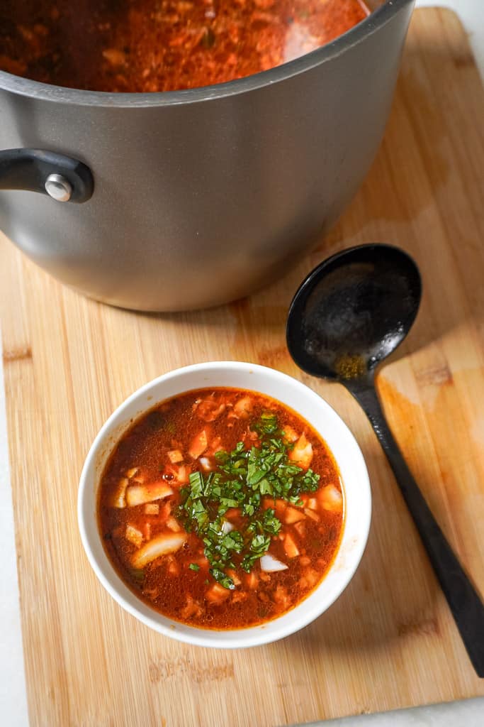 a bowl of birria consome beside a large pot containing more and a ladle spoon beside it