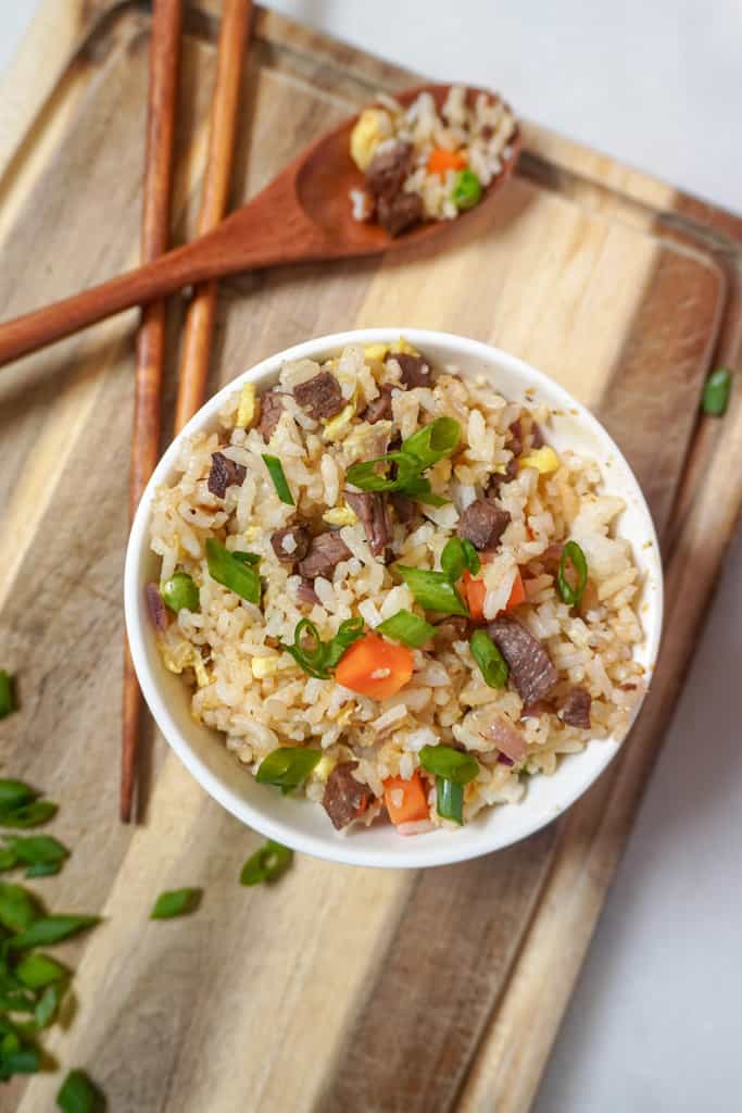 a bowl of steak fried rice on a cutting board beside chopsticks and a wooden spoon