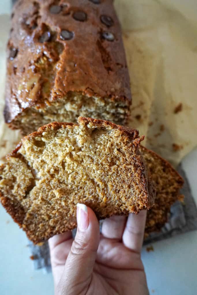 a hand holding a slice of banana bread with the rest of the loaf in the background