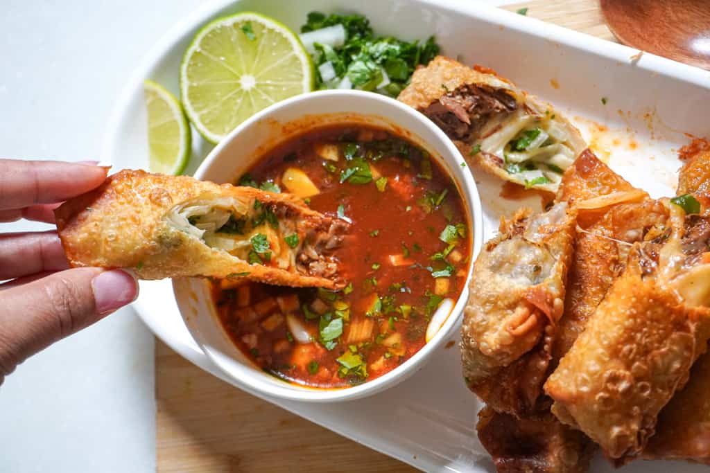 a birria egg roll being dipped into a birria sauce, on a platter with other egg rolls