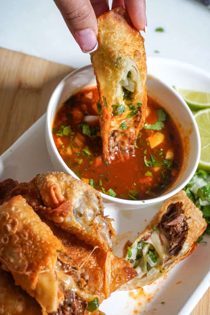 a hand dipping an egg roll in birria sauce, on a platter with more egg rolls