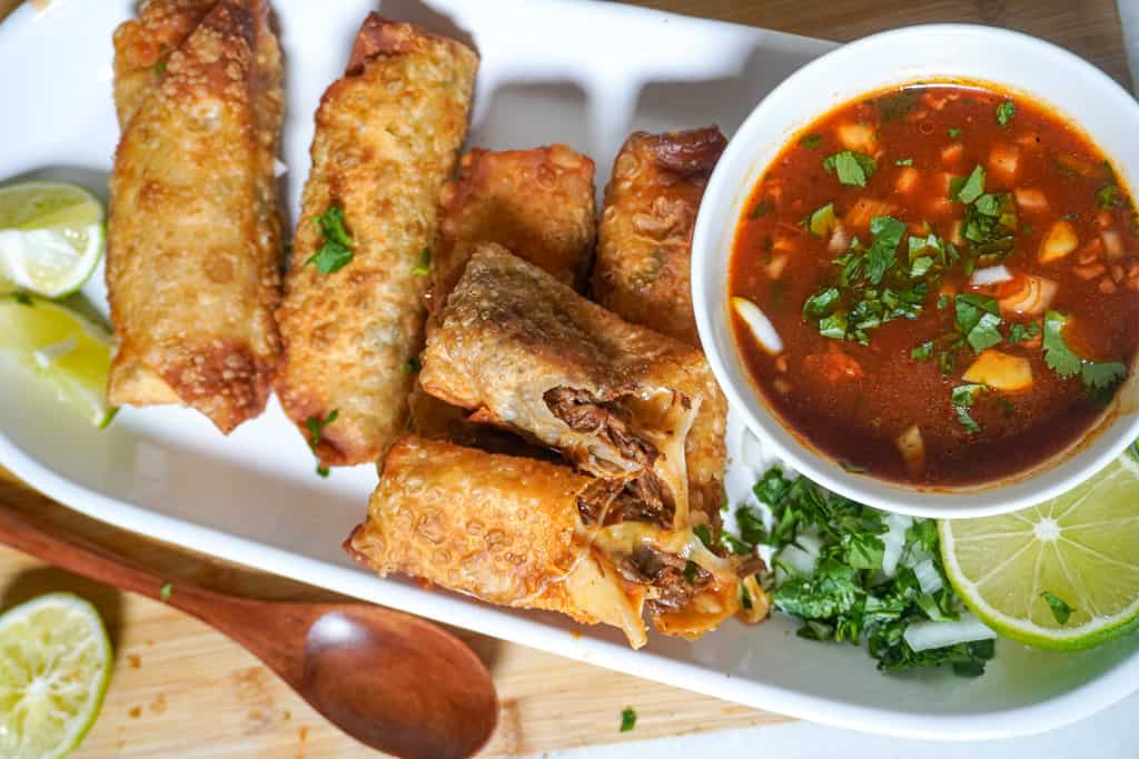 a platter of birria egg rolls with a saucer of birria