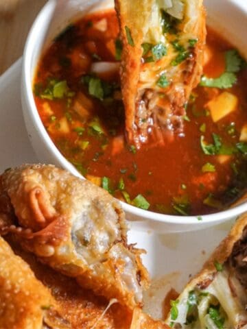 a hand dipping an egg roll in birria sauce, on a platter with more egg rolls