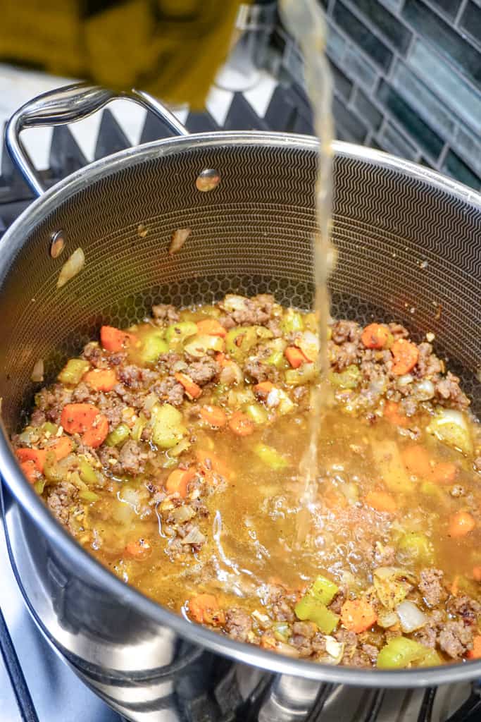 broth being poured into a large stockpot with chopped veggies and sausage