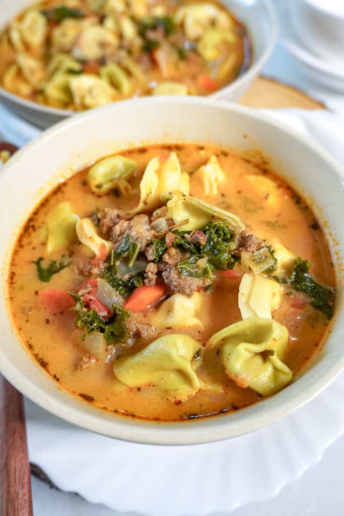 a bowl of sausage tortellini soup with another bowl in the background, out of focus