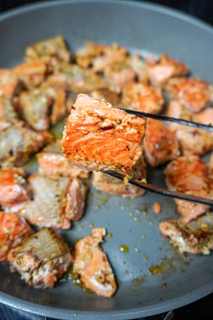 tongs holding a piece of lemon pepper salmon over a pan of more