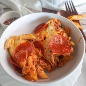 A serving of pizza pasta topped with melted cheese and pepperoni slices in a white bowl, with utensils and seasonings in the background.