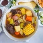 a bowl of beef and vegetable soup with a side of lime wedges and Mexican rice on a table setting