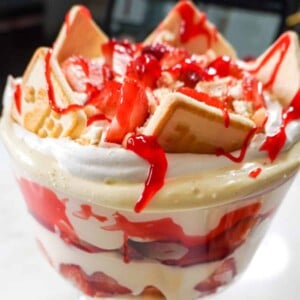 A close-up of a Strawberry Banana Pudding trifle, featuring creamy layers of pudding, fresh strawberries, bananas, Chessman cookies, and a drizzle of strawberry glaze on top.