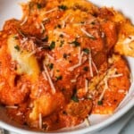 A close-up of crockpot lasagna served in a white bowl, covered in marinara sauce, melted cheese, and topped with grated Parmesan and fresh herbs.