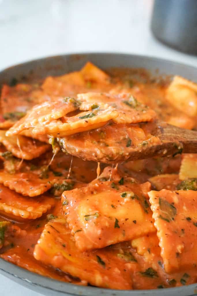 Cheese ravioli being scooped from a skillet filled with creamy tomato sauce and herbs.