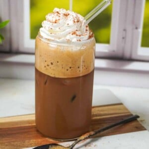 A tall glass of brown sugar shaken espresso topped with whipped cream and cinnamon, served with a glass straw and a spoon of foamy espresso mixture on the side.