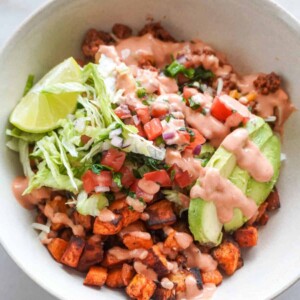 A taco bowl filled with roasted sweet potatoes, ground beef, avocado slices, shredded lettuce, pico de gallo, and a drizzle of creamy pink sauce, garnished with lime.